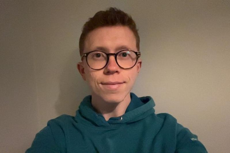 head and shoulders image of a smiling young man with short, red, curly hair wearing dark-rimmed glasses and a dark blue hoody
